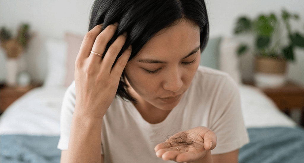 : A woman in a white t-shirt looking down at a small clump of fallen hair strands held in the palm of her hand.