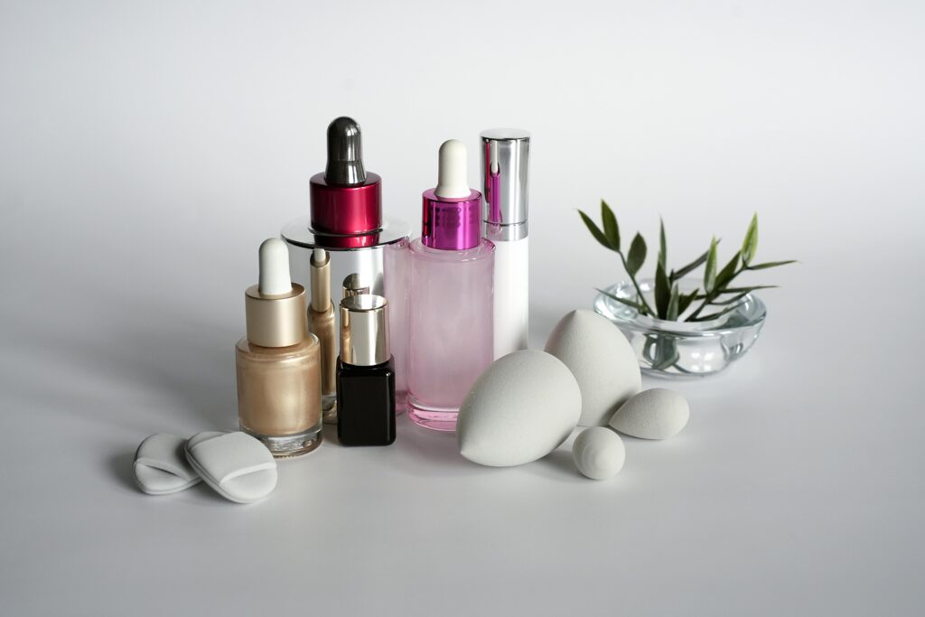 A clean, minimalist arrangement of beauty products on a white surface. The collection includes various dropper bottles with gold and metallic pink caps, a white pump bottle, and several grey teardrop-shaped makeup sponges, accented by a small green plant in a glass bowl. acne