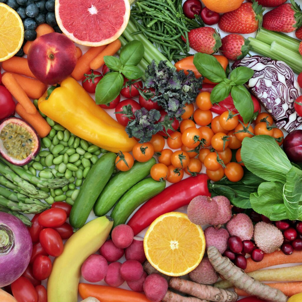 A top-down, colorful flat lay of various fresh produce including bell peppers, tomatoes, carrots, citrus fruits, leafy greens, radishes, and berries.
