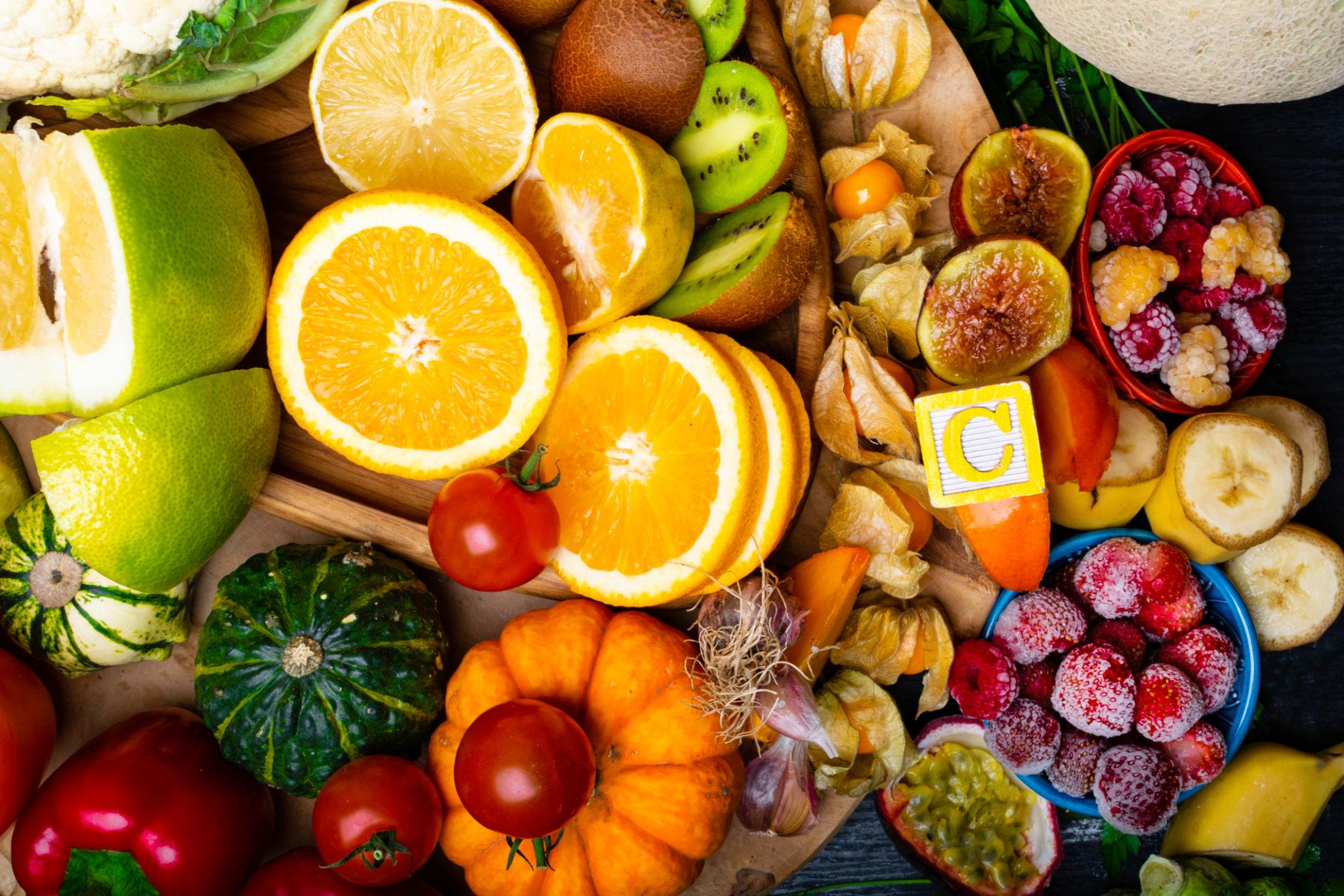 A vibrant, high-angle close-up of various whole and sliced produce, including oranges, kiwi, raspberries, and tomatoes, surrounding a yellow block labeled with the letter C to represent an antioxidant vitamin source.