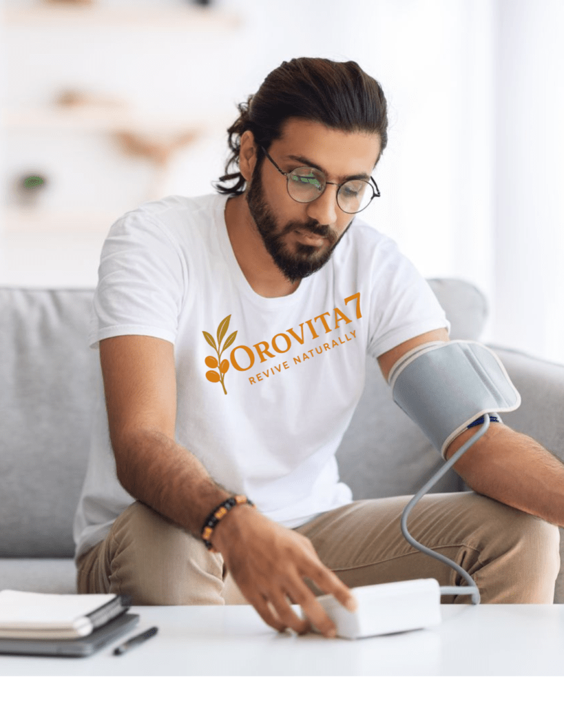 A young man in an OroVita7 "Revive Naturally" t-shirt sitting on a sofa and using a digital blood pressure monitor on his arm.