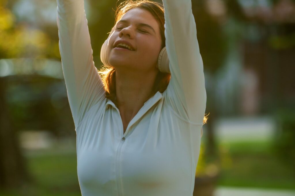 A young woman in white athletic gear and headphones stretching her arms high above her head with her eyes closed, enjoying the warm golden sunlight in an outdoor park setting.