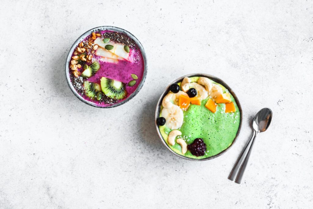 Top-down view of two colorful smoothie bowls on a white textured surface; one bright pink topped with kiwi and chia seeds, the other bright green topped with mango, banana, and cashews.
