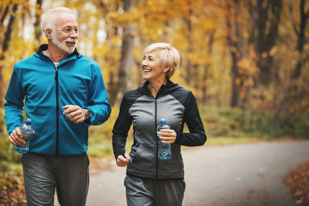 A happy senior man and woman in athletic wear jogging together on a paved path through a forest with golden autumn leaves, both holding water bottles and smiling. boost