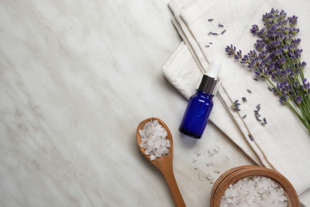 A top-down view of Botanical wellness ingredients featuring fresh lavender sprigs, a blue glass dropper bottle, and coarse white sea salt in a wooden spoon on a marble surface. Botanical