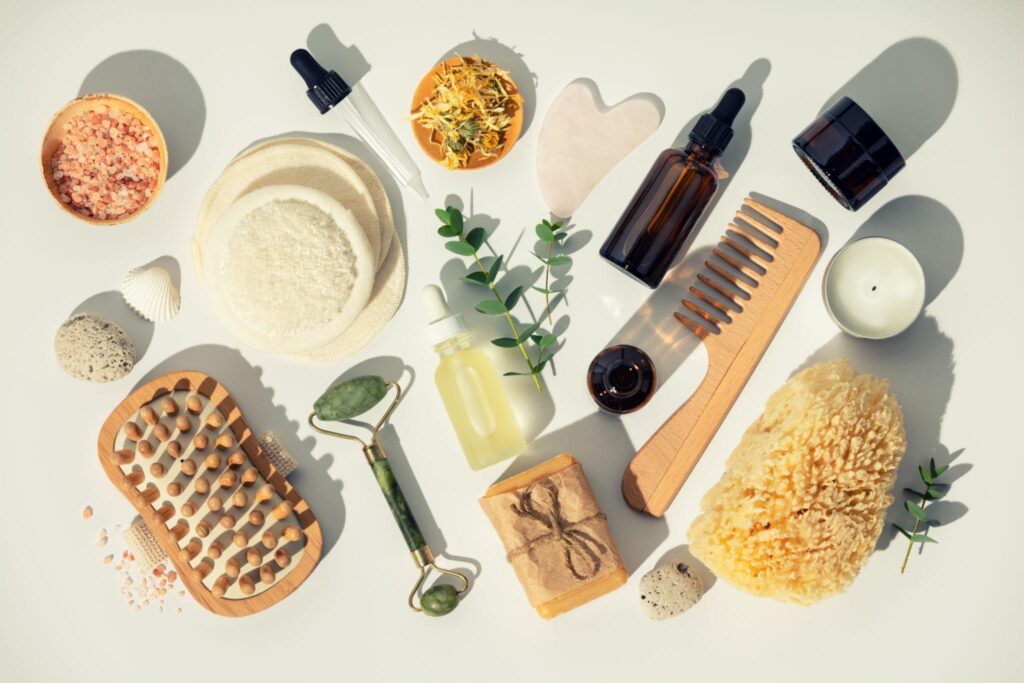 A collection of botanical wellness items featuring a jade roller, gua sha tool, wooden hairbrush, dried flowers, and various amber glass tincture bottles casting soft shadows.