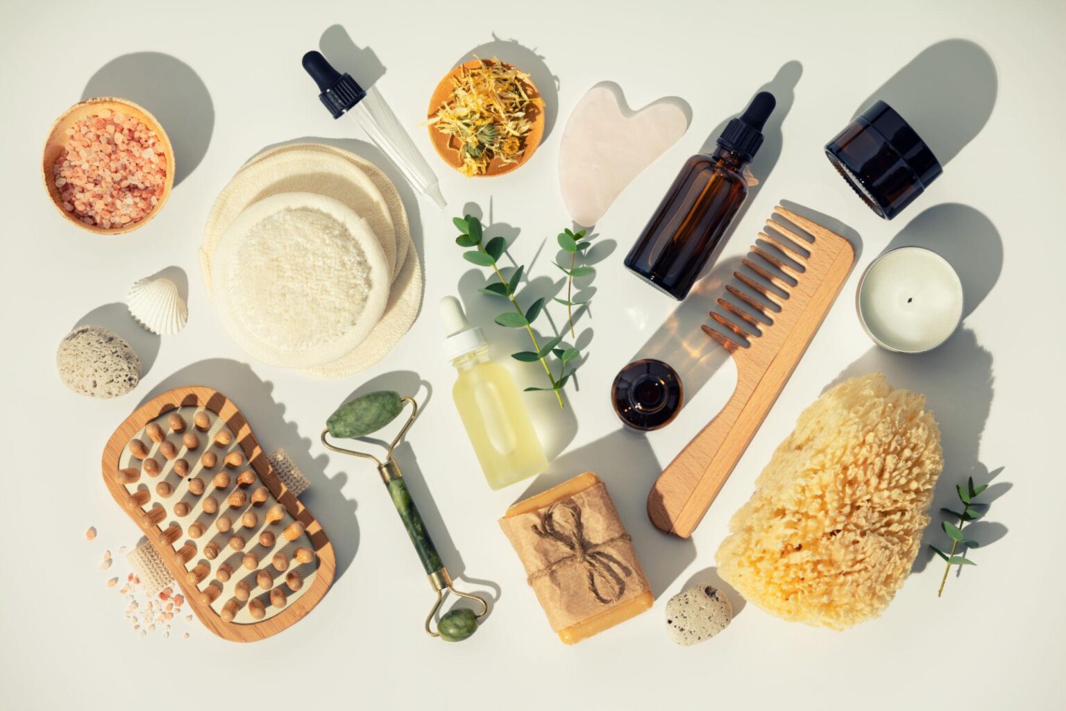 A collection of botanical wellness items featuring a jade roller, gua sha tool, wooden hairbrush, dried flowers, and various amber glass tincture bottles casting soft shadows.