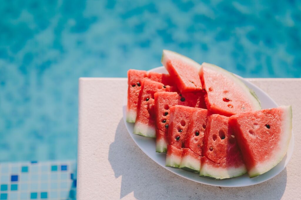 A high-angle, close-up shot of a white plate filled with several triangular slices of ripe, red watermelon with black seeds. The plate sits on the edge of a light-colored stone pool deck. In the background, the bright blue, shimmering water of a swimming pool is softly blurred, creating a sunny and refreshing summer atmosphere.