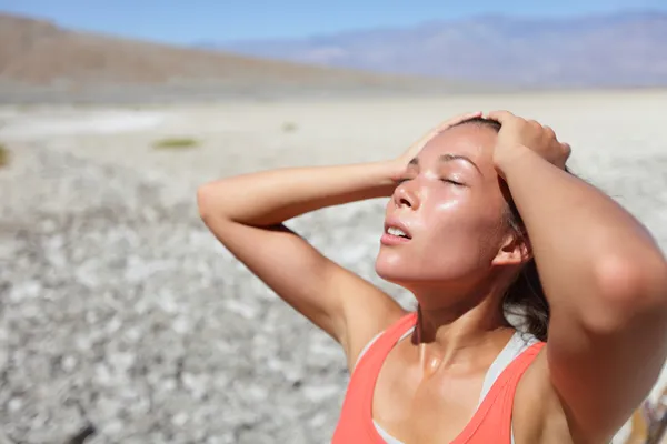 A woman in an orange tank top stands in a dry, rocky desert landscape, squinting with her hands on her head as she reacts to intense heat and sun exposure.
