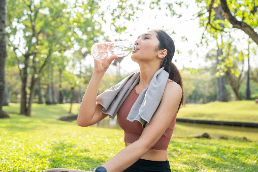 A woman in athletic wear sits on green grass in a sunlit park, drinking water from a clear plastic bottle with a grey towel draped around her neck.