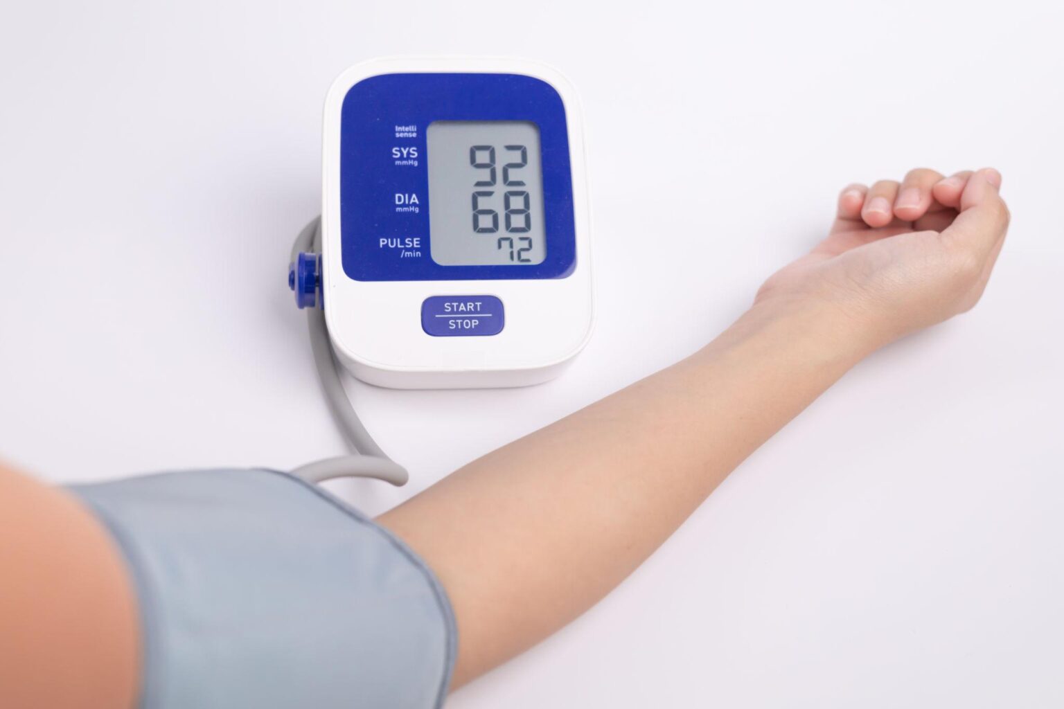 A high-angle, close-up shot of a person's arm with a grey blood pressure cuff attached, positioned next to a digital monitor on a white surface. The blue and white monitor displays a systolic reading of 92, a diastolic reading of 68, and a pulse of 72.