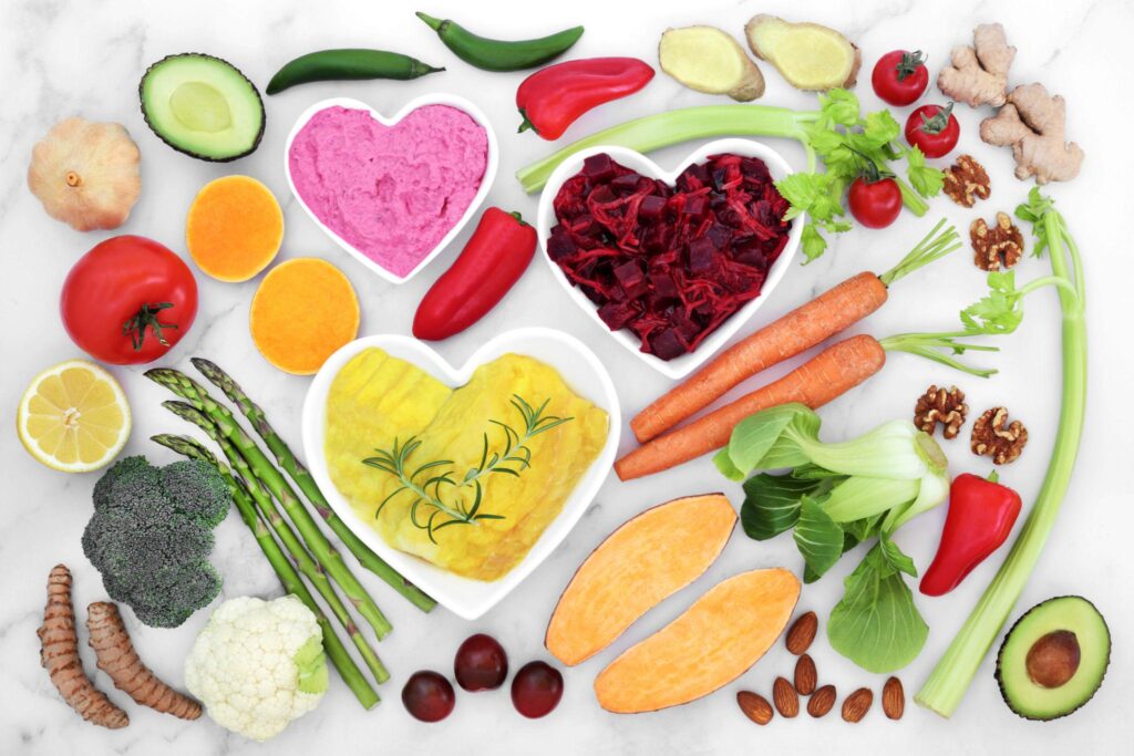 A bright, overhead shot of diverse fresh produce and healthy ingredients on a white marble background. Three heart-shaped bowls contain beet salad, hummus, and a yellow turmeric-based dip. The arrangement includes asparagus, cauliflower, turmeric root, ginger, bell peppers, leafy greens, avocado halves, and whole walnuts, symbolizing a balanced and healthy diet.