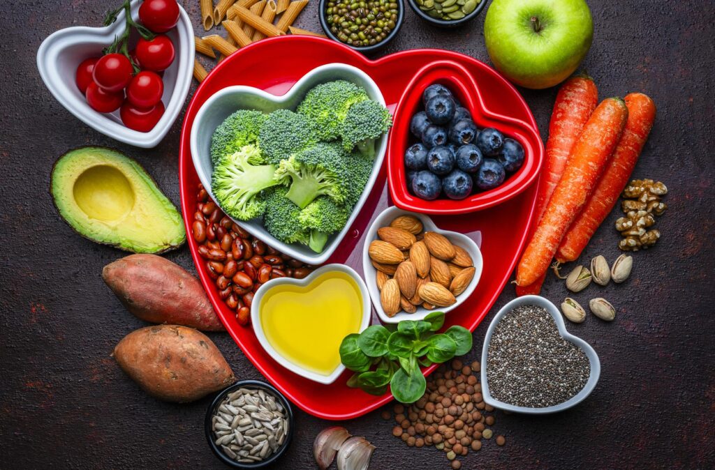 A top-down view of a vibrant collection of heart-healthy foods centered around a large red heart-shaped tray. The tray holds bowls of broccoli, blueberries, almonds, and olive oil. Surrounding the tray on a dark, rustic surface are ingredients like avocado, sweet potatoes, carrots, tomatoes, chia seeds, lentils, walnuts, and a green apple.