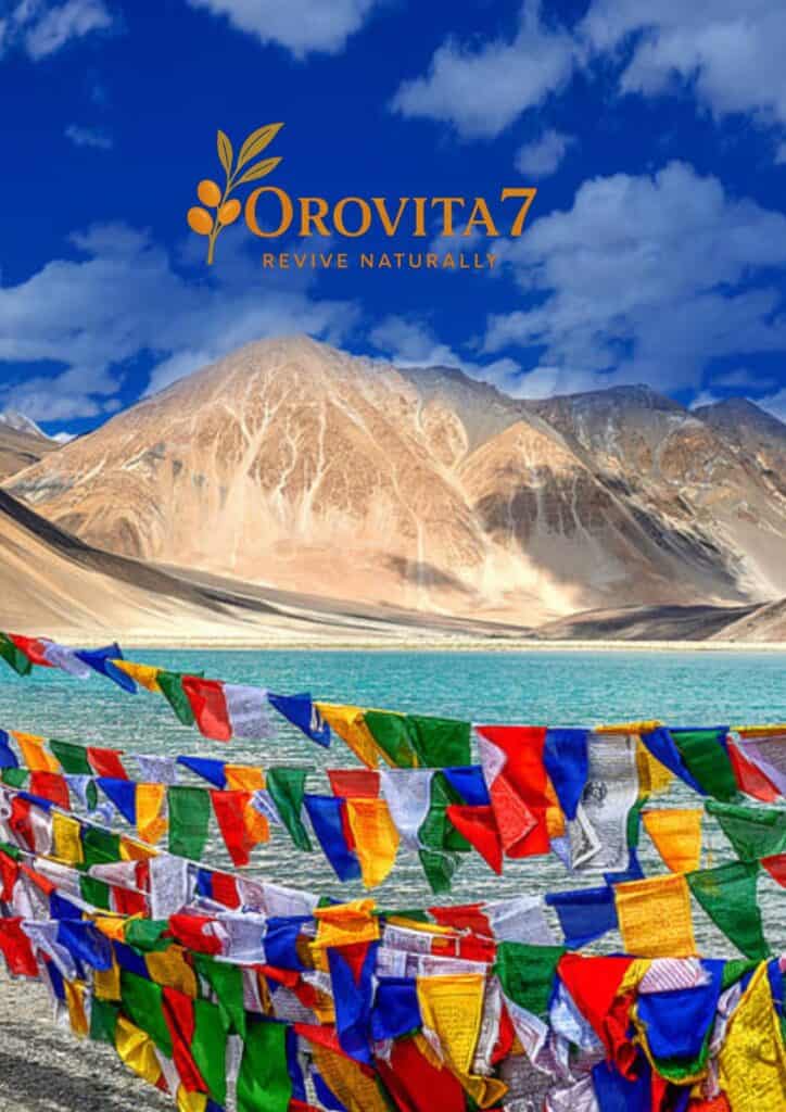 Colorful Tibetan prayer flags fluttering in front of a turquoise high-altitude lake and barren mountains in Ladakh, with the OroVita7 Revive Naturally logo overlaid.