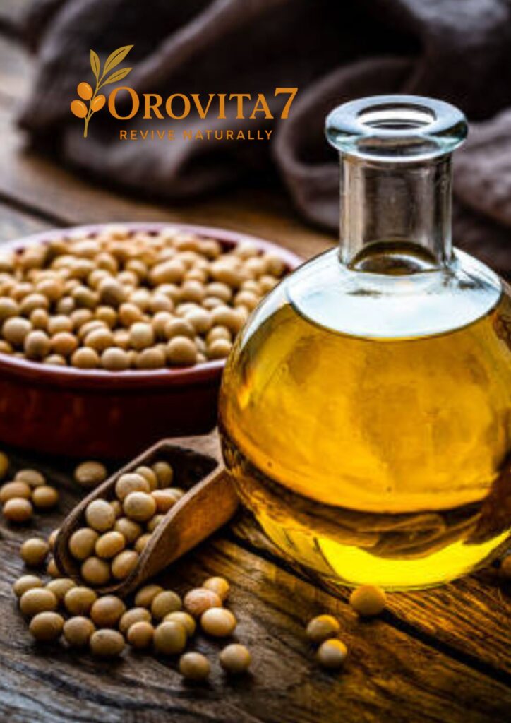 A round glass carafe filled with amber-colored soy oil sitting on a wooden table next to a bowl and scoop of raw soybeans.