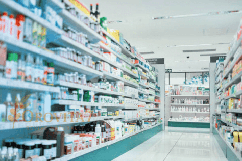 A wide-angle view of a bright, modern pharmacy with rows of white shelves stocked with various healthcare products and medications. The OROVITA7 "Revive Naturally" logo is watermarked in the lower-left corner.