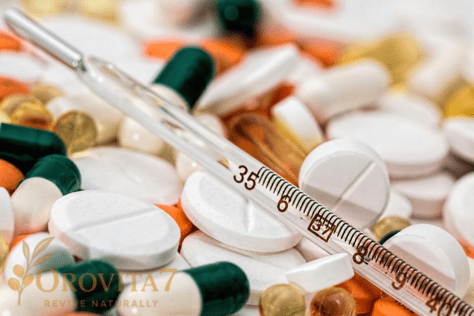 A macro shot showing a glass medical thermometer resting on a scattered pile of white, orange, and green pills and capsules. The OROVITA7 logo is visible in the bottom-left corner.