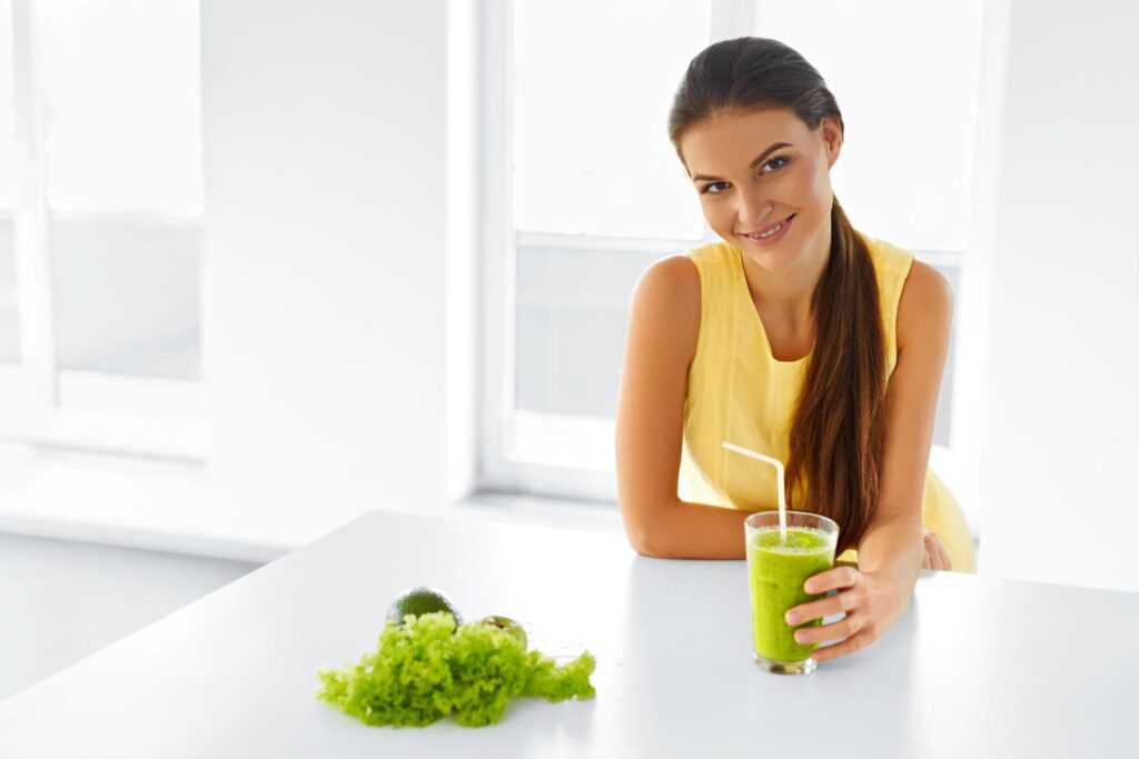 A smiling woman in a yellow top holding a glass of green vegetable smoothie at a white table with fresh greens and an avocado. prevention