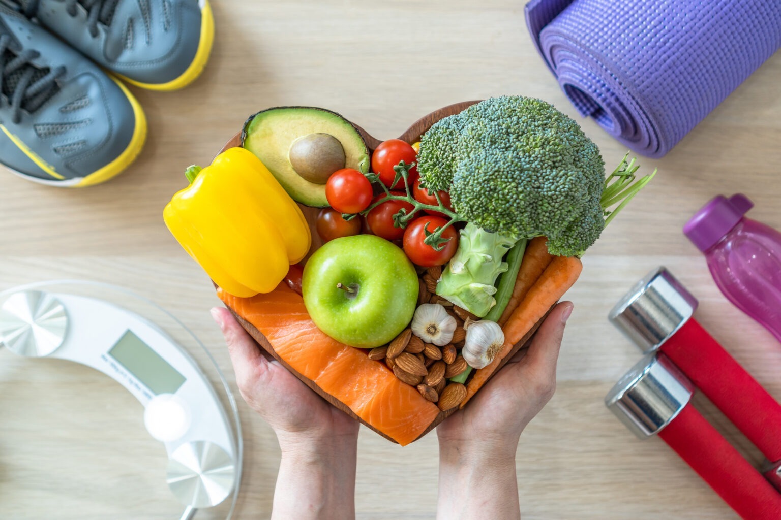 Overhead view of hands holding a heart-shaped wooden bowl filled with salmon, broccoli, avocado, tomatoes, apples, and almonds, surrounded by fitness equipment like dumbbells and a yoga mat.