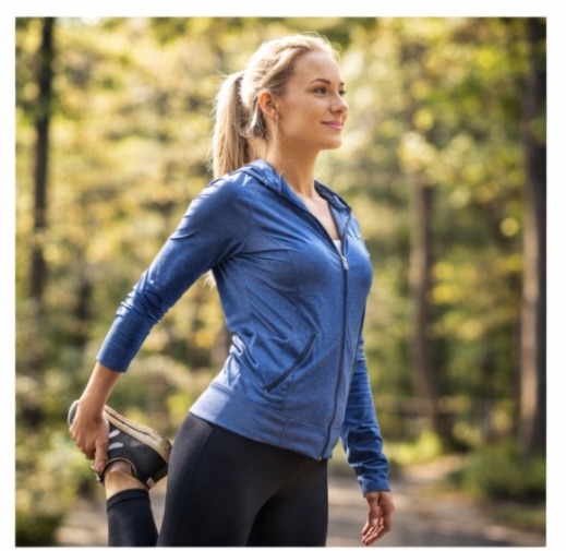 A blonde woman in a blue athletic zip-up hoodie and black leggings performing a standing quad stretch in a sunlit park or wooded area.
