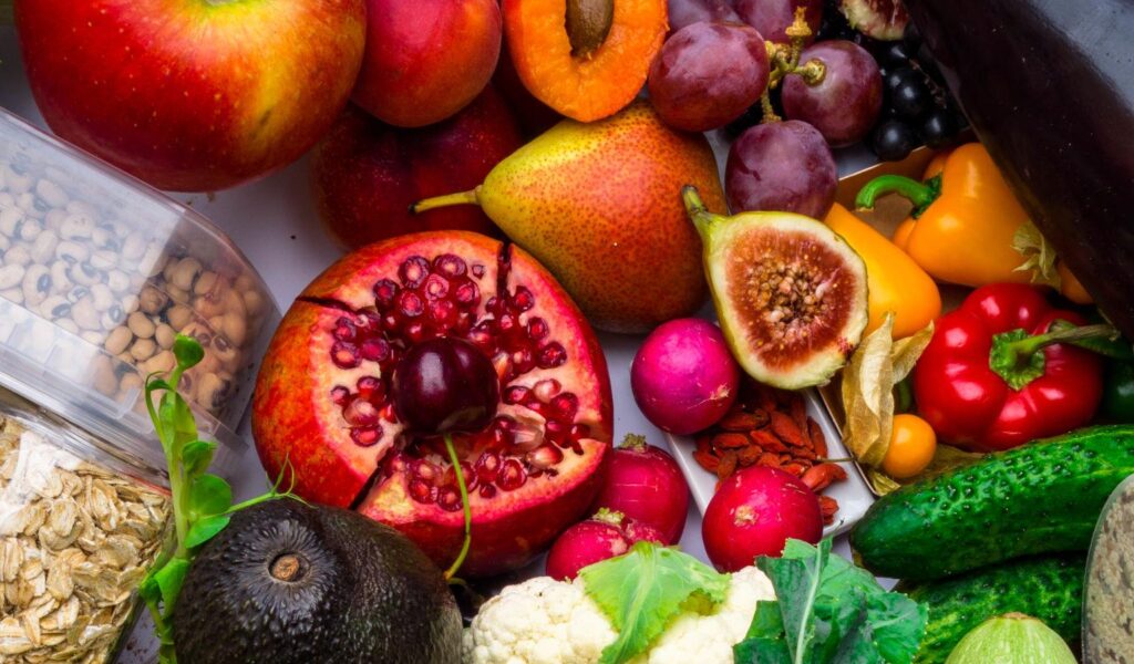 A top-down, crowded arrangement of various fresh foods including a large red apple, a halved pomegranate, a dark avocado, cauliflower, leafy green sprouts, and cucumbers. Containers of oats, legumes, and chia seeds are tucked among the produce, creating a colorful display of a plant-based diet.