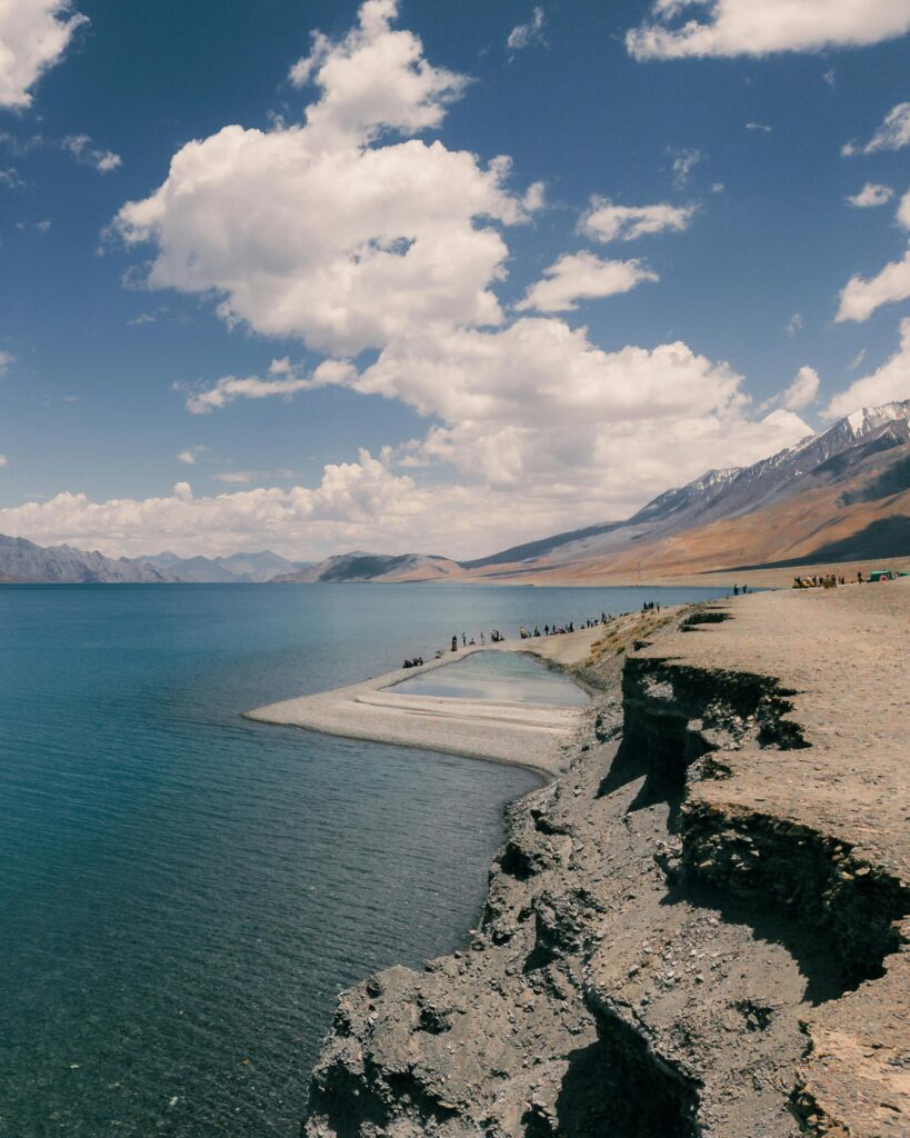 “Scenic Pangong Lake in Ladakh with blue water, rocky shoreline, and distant Himalayan mountains under cloudy sky”