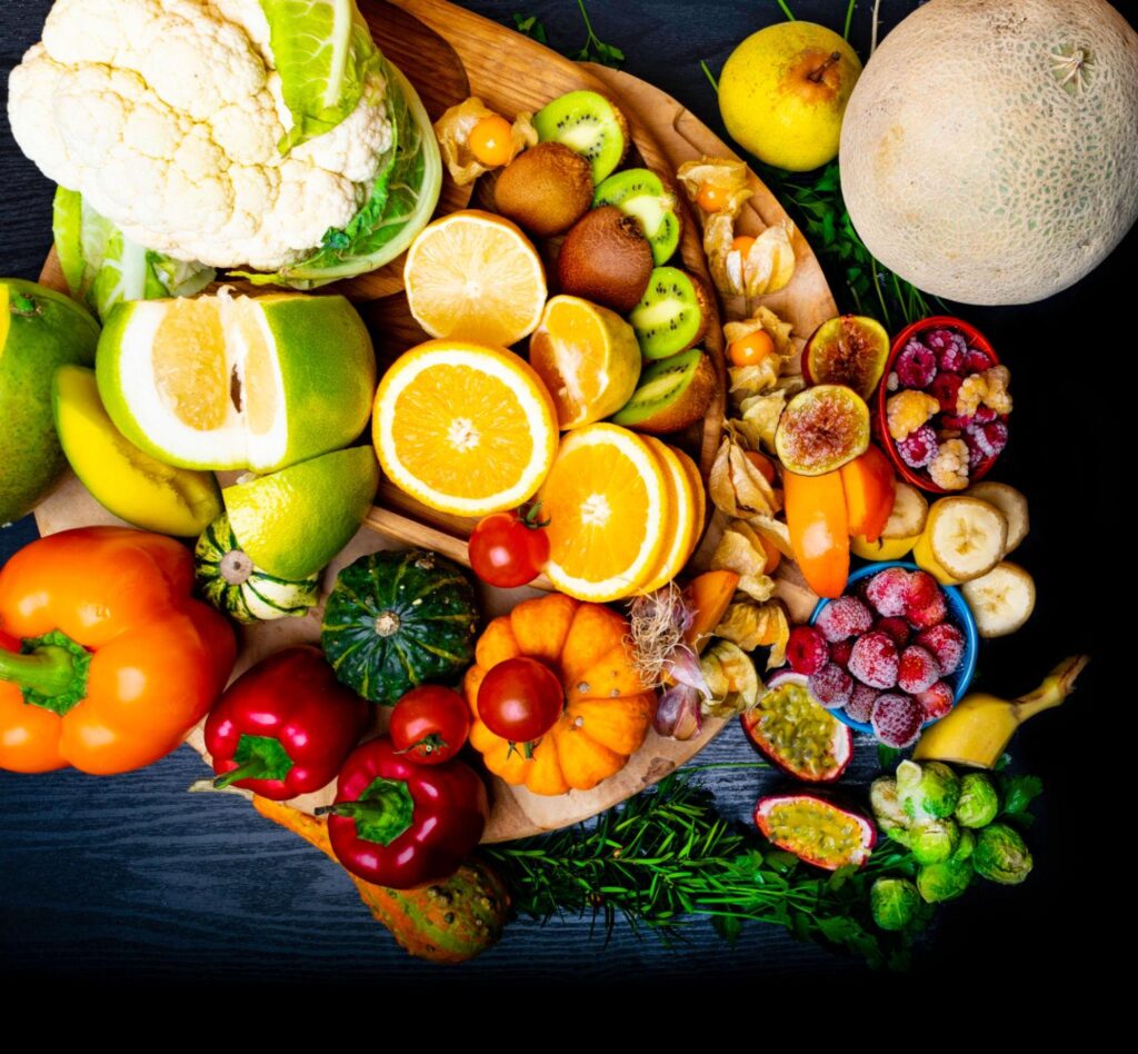 A colorful collection of sliced oranges, kiwi, cauliflower, peppers, passion fruit, raspberries, and melons arranged on a dark background.