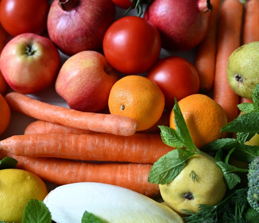 A dense arrangement of bright red tomatoes, pomegranates, orange carrots, apples, lemons, and fresh mint leaves.