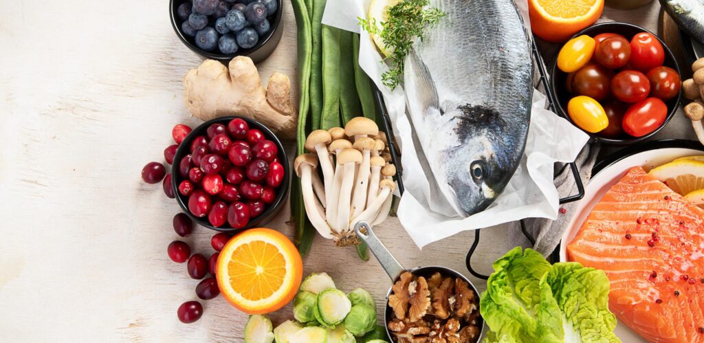 A flat-lay photograph of various healthy food groups on a white wooden surface. It features a whole raw fish, a salmon fillet, blueberries, cranberries, ginger root, mushrooms, walnuts, leafy greens, and citrus fruits. vitamin deficiency
