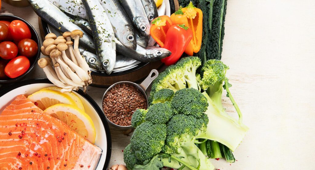 A close-up shot of raw salmon, sardines, broccoli, mushrooms, and bell peppers. These nutrient-dense ingredients are shown as essential dietary choices for improving weak immunity.