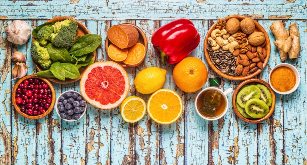 A top-down view of various whole foods on a rustic blue wooden background. The spread includes citrus fruits, berries, nuts, seeds, and leafy greens, all known for supporting those with weak immunity.